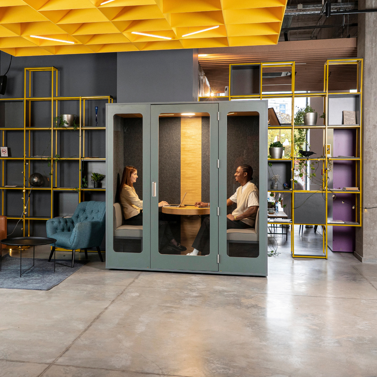 A modern office interior with two people seated inside a large enclosed meeting pod with glass doors. The pod is muted green-gray with a wood-look back wall and warm ambient lighting.
