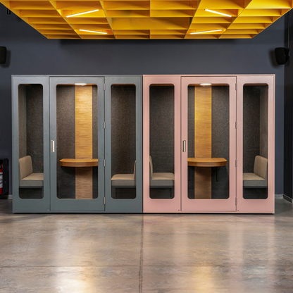 Two modern acoustic meeting pods with glass doors, one in slate gray and one in blush pink, shown side by side in a contemporary office space with a concrete floor and a yellow ceiling grid.