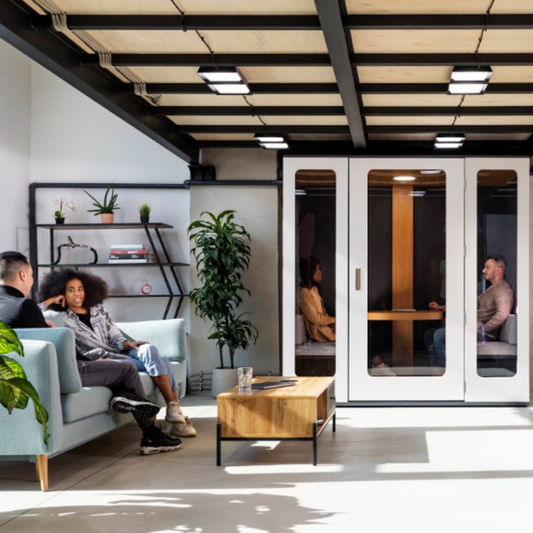 A modern office interior with a light blue sofa, a wooden coffee table, shelving, and a glass-walled privacy pod where several people are seated and talking.